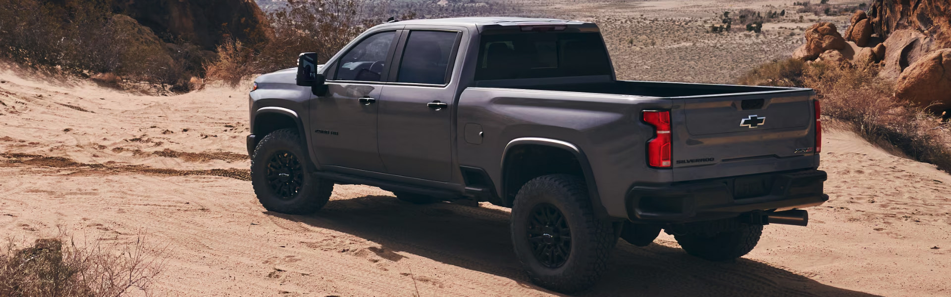A gray 2025 Chevrolet Silverado 2500 pickup truck parked on a sandy desert trail, showing its rugged design and off-road stance.
