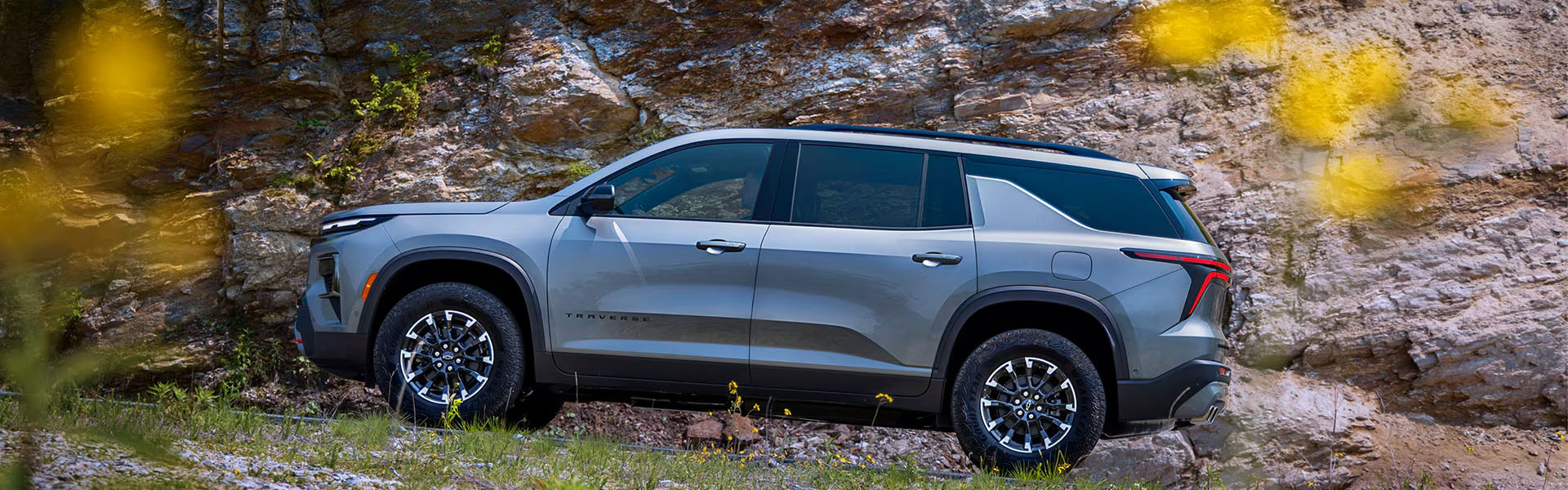 Side view of a gray 2026 Chevrolet Traverse with black wheels parked along a rocky roadside surrounded by natural landscape.