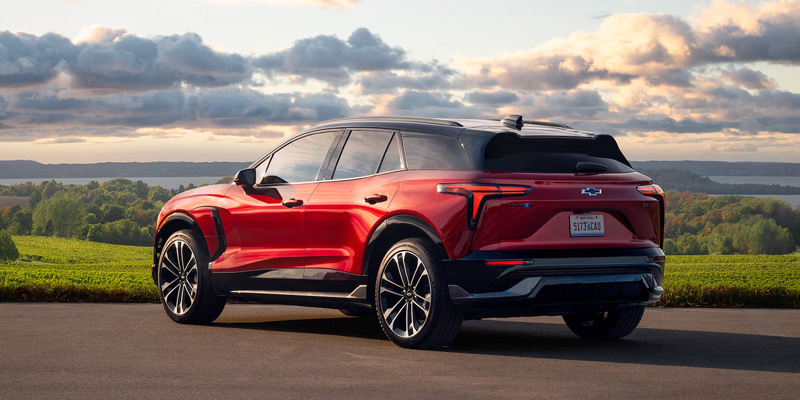 A sleek red Chevrolet Blazer EV is parked on a paved road, overlooking a lush green landscape under a cloudy sky at sunset. The scene conveys tranquility and innovation.