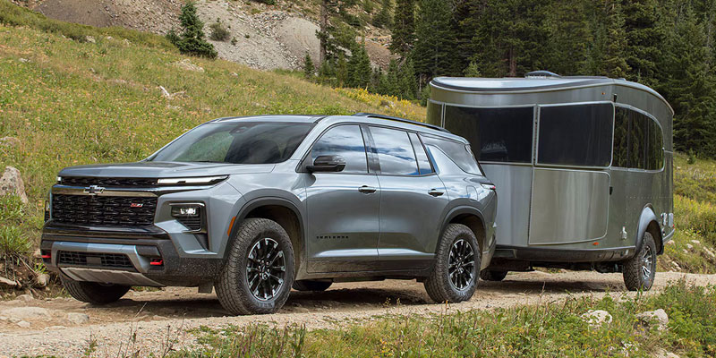 A gray Chevrolet Traverse pulls a sleek, modern trailer on a rustic dirt path, surrounded by lush green trees and hills, evoking a sense of adventure and exploration.
